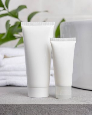 Paket maketi, beyaz sıkılmış krema tüpleri yeşil yapraklı dal, leğen ve beyaz banyo havluları banyodaki gri taş tezgahın üzerinde. Doğal bitkisel kozmetik