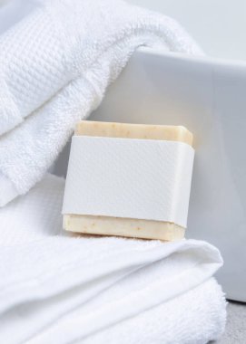 Havlunun yanındaki beyaz banyo havlularının üzerinde boş etiketli krem sabun kalıbı, yakın çekim, ambalaj modeli. Doğal el yapımı hijyen ürünü. Temiz tarafsız banyo, yumuşak ışık.