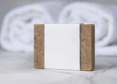 Beyaz banyo havlularının yanında boş kağıt etiketli kahverengi, katı sabun kalıbı. Tarafsız arkaplanda fotokopi alanı olan markasız ürün. Kişisel bakım bağlamı