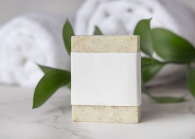 Beyaz banyo havlularının yanında açık bej, katıksız sabun kalıbı, boş kağıt etiket ve yeşil yapraklar. Tarafsız arkaplanda fotokopi alanı olan markasız ürün. Kişisel bakım ve kaplıca içeriği