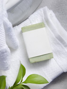Yeşil sabun kalıbının ambalajı. Beyaz banyo havlusunun üzerinde boş etiket ve gri taş tezgahın üstünde yeşil yapraklar. Doğal el yapımı bitkisel hijyen ürünü. Tarafsız tuvalet
