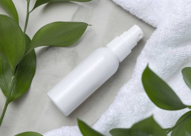 Beyaz banyo havlusunun yanında boş beyaz sprey şişesi ve mermerin üzerinde yeşil yapraklar. Kopya alanı ile markasız kozmetik paketleme. Tarafsız tuvalet sahnesi, üst manzara. Damgalama şablonu