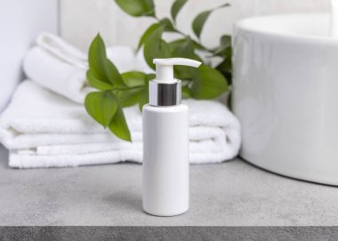 Paket maketi, beyaz boş bir pompa krem şişesi yeşil yapraklı dal, leğen ve beyaz banyo havlularına karşı banyodaki gri taş tezgahın üstünde. Doğal bitkisel kozmetik