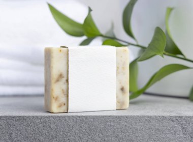 Yeşil yapraklı, beyaz banyo havluları ve gri taş tezgahın üzerinde boş etiketli krem sabun kalıbı kapatılıyor, ambalaj modeli yapılıyor. Doğal el yapımı bitkisel hijyen ürünü. Tarafsız tuvalet