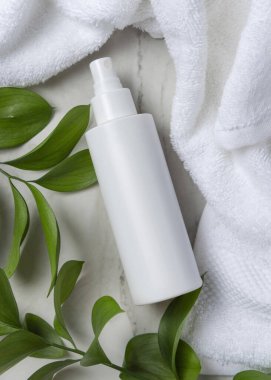 Beyaz banyo havlusunun yanında boş beyaz sprey şişesi ve mermerin üzerinde yeşil yapraklar. Kopya alanı ile markasız kozmetik paketleme. Tarafsız tuvalet sahnesi, üst manzara. Damgalama şablonu