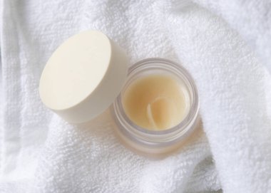 Beyaz havlu üzerinde boş kapaklı dudak kremi kavanozu, üst manzara. Tarafsız arkaplanda fotokopi alanı ile markasız kozmetik ambalaj. Spa otel banyosu içeriği