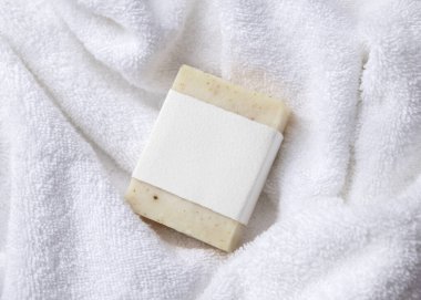 Beyaz banyo terli havlularının üzerinde boş kağıt etiketli bej, katı sabun kalıbının bir kopyası. Tarafsız arkaplanda, fotokopi alanı ile markasız paketleme, üst görünüm. Kişisel bakım bağlamı