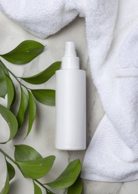 Beyaz banyo havlusunun yanında boş beyaz sprey şişesi ve mermerin üzerinde yeşil yapraklar. Kopya alanı ile markasız kozmetik paketleme. Tarafsız tuvalet sahnesi, üst manzara. Damgalama şablonu