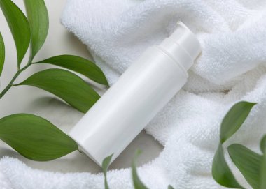 Beyaz terry havlunun üzerindeki boş beyaz pompa şişesinin maketi taşın üzerindeki yeşil yaprakların yanında, yakın plan. Kopya alanı ile markasız kozmetik paketleme. Cilt ürünüyle tarafsız banyo sahnesi