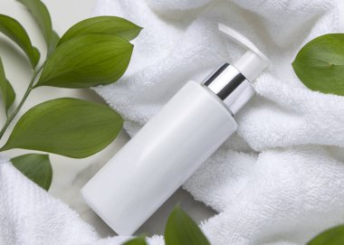 Beyaz Terry havlusunun yanında boş beyaz pompa şişesi ve taşın üstünde yeşil yapraklar, üst manzara. Gümüş yakalı ve fotokopi alanlı markasız kozmetik ambalajlar. Cilt ürünleri