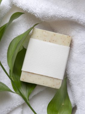 Beyaz banyo havlularının üzerindeki yeşil yaprakların yanında boş kağıt etiketli bej, katı sabun kalıbının bir kopyası. Tarafsız arkaplanda fotokopi alanı olan markasız ürün. Kişisel bakım ve kaplıca içeriği