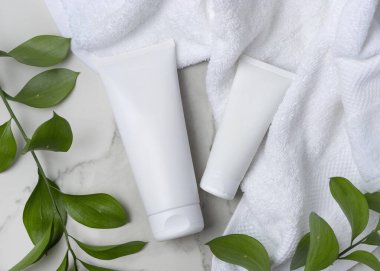 Beyaz havlunun yanında beyaz krema tüpleri ve taşın üstünde yeşil yapraklar. Kopya alanı ile markasız kozmetik paketleme. Cilt ürünüyle tarafsız banyo sahnesi