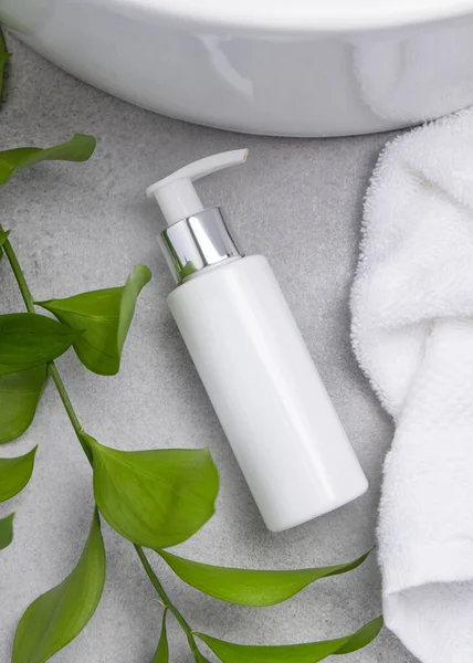 Paket maketi, beyaz boş bir pompa krem şişesi yeşil yapraklı dal, leğen ve beyaz banyo havlularıyla banyo manzaralı gri taş tezgahta. Doğal bitkisel kozmetik
