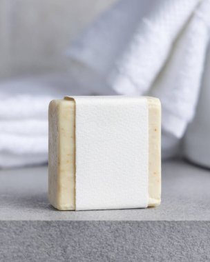 Beyaz banyo havlularının yanında boş etiketli krem sabun barı ve gri taş tezgahın üzerinde leğen, yakın plan, ambalaj modeli. Doğal el yapımı hijyen ürünü. Temiz tarafsız banyo içi.
