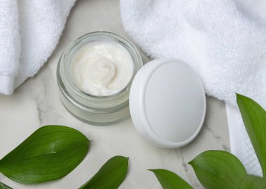 Beyaz havlunun yanında boş kapağı ve taşın üstünde yeşil yaprakları olan açık yüz kremi kavanozunun bir kopyası. Kopya alanı ile markasız kozmetik paketleme. Cilt ürünüyle tarafsız banyo sahnesi