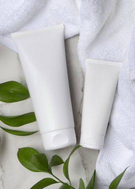 Beyaz havlunun yanında boş beyaz krema tüpleri ve taşın üstünde yeşil yapraklar. Kopya alanı ile markasız kozmetik paketleme. Cilt ürünüyle tarafsız banyo sahnesi