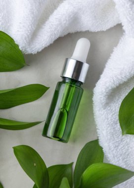 Beyaz Terry Havlu 'nun yanında kapalı yeşil cam damlatıcı şişesi ve yapraklar, üst manzara, maket. Botanik tarzda spa ve otel cilt ürünleri için markasız kozmetik ambalajlama