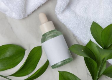 Beyaz banyo havlusunun yanında kapalı cam damlatıcı şişesi ve yeşil yapraklar üst manzara. Etiketli markasız kozmetik ambalaj modeli. Cilt bakımı - serum ya da öz, kopyalama alanı olan tarafsız banyo sahnesi