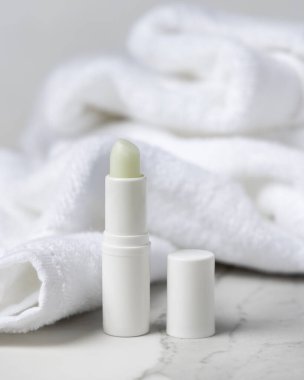 Beyaz banyo havlusunun yanında kapağı olan boş bir dudak kremi çubuğu. Gri taş tezgahında boş etiket alanı olan markasız kozmetik ambalajlama, nötr arkaplan ve boşluk kopyalama