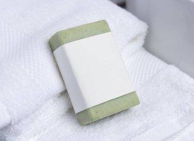 Havlunun yanındaki beyaz banyo havlularının üzerinde boş etiketi olan yeşil sabun kalıbı, yakın çekim, ambalaj modeli. Doğal el yapımı hijyen ürünü. Temiz tarafsız banyo, yumuşak ışık.