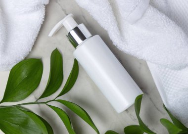 Beyaz Terry havlusunun yanında boş beyaz pompa şişesi ve taşın üstünde yeşil yapraklar, üst manzara. Gümüş yakalı ve fotokopi alanlı markasız kozmetik ambalajlar. Cilt ürünleri