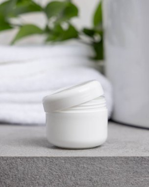 Beyaz havlunun yanında boş tarafı ve taşın üstünde yeşil yaprakları olan açık yüz kremi kavanozunun bir kopyası. Kopya alanı ile markasız kozmetik paketleme. Cilt ürünüyle tarafsız banyo sahnesi