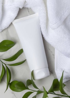 Beyaz Terry havlusunun yanında beyaz krema tüpü ve taşın üstünde yeşil yapraklar. Kopya alanı ile markasız kozmetik paketleme. Cilt ürünüyle tarafsız banyo sahnesi