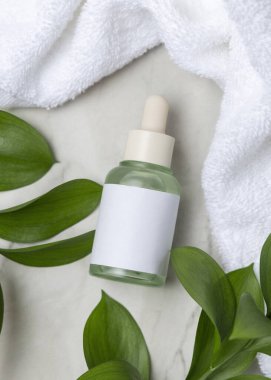 Beyaz banyo havlusunun yanında kapalı cam damlatıcı şişesi ve taşın üstünde yeşil yapraklar. Etiketli markasız kozmetik ambalaj modeli. Tarafsız banyo sahnesinde cilt bakımı, kopyalama alanı