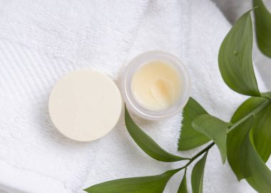 Yeşil yaprakların yanında beyaz havlu üzerinde doğal bitkisel merhem bulunan açık merhem kavanozu. Boş kapaklı kozmetik ambalaj ve markalaşma için kopyalama alanı. Dudak bakım ürünü