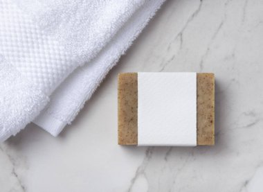 Beyaz banyo terry havlularının yanında boş kağıt etiketli kahverengi, katı sabun kalıbının bir kopyası. Tarafsız arka planda markasız ürün, kopya alanı, üst görünüm. Kişisel bakım bağlamı