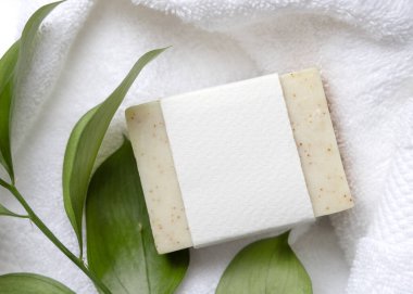 Beyaz banyo havlularının üzerindeki yeşil yaprakların yanında boş kağıt etiketli bej, katı sabun kalıbının bir kopyası. Tarafsız arkaplanda fotokopi alanı olan markasız ürün. Kişisel bakım ve kaplıca içeriği