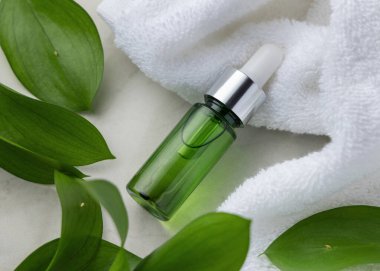 Beyaz Terry Havlu 'nun yanında kapalı yeşil cam damlatıcı şişesi ve yapraklar, taşa yakın, maket. Botanik tarzda spa ve otel cilt ürünleri için markasız kozmetik ambalajlama