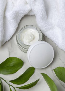 Beyaz havlunun yanında boş kapağı ve taşın üstünde yeşil yaprakları olan açık yüz kremi kavanozunun bir kopyası. Kopya alanı ile markasız kozmetik paketleme. Cilt ürünüyle tarafsız banyo sahnesi