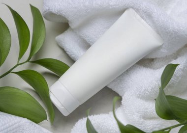 Beyaz havlunun yanında boş beyaz krema tüpü ve taşın üstünde yeşil yapraklar. Kopya alanı ile markasız kozmetik paketleme. Cilt ürünüyle tarafsız banyo sahnesi