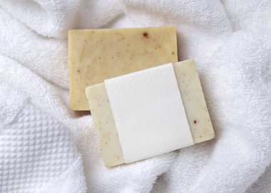 Beyaz banyo terli havlularının üzerinde boş kağıt etiketli bej, katı sabun kalıbının bir kopyası. Tarafsız arkaplanda, fotokopi alanı ile markasız paketleme, üst görünüm. Kişisel bakım bağlamı