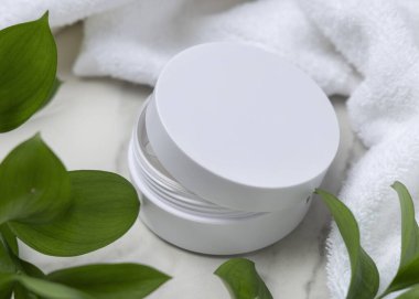 Beyaz havlu ve yeşil yaprakların yanında boş kapaklı kozmetik krem kavanozu açıldı. Yüz bakımı ve spa için cilt bakımı ürünü