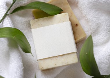 Beyaz banyo havlularının üzerindeki yeşil yaprakların yanında boş kağıt etiketli bej, katı sabun kalıbının bir kopyası. Tarafsız arkaplanda fotokopi alanı olan markasız ürün. Kişisel bakım ve kaplıca içeriği