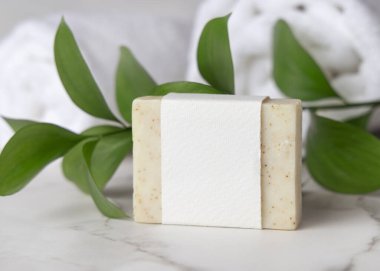 Beyaz banyo havlularının yanında açık bej, katıksız sabun kalıbı, boş kağıt etiket ve yeşil yapraklar. Tarafsız arkaplanda fotokopi alanı olan markasız ürün. Kişisel bakım ve kaplıca içeriği