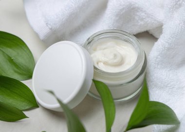 Beyaz havlunun yanında boş kapağı ve taşın üstünde yeşil yaprakları olan açık yüz kremi kavanozunun bir kopyası. Kopya alanı ile markasız kozmetik paketleme. Cilt ürünüyle tarafsız banyo sahnesi