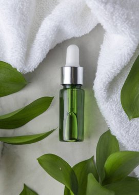 Beyaz havlu ve yaprakların yanında kapalı yeşil cam damlatıcı şişesi, taşın üstünde üst manzara, maket. Botanik tarzda spa ve otel cilt ürünleri için markasız kozmetik ambalajlama