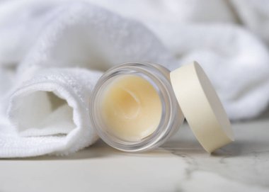 Dudak kremi kavanozunun kapağı beyaz banyo havlusuyla açılmış. Tarafsız arkaplanda fotokopi alanı ile markasız kozmetik ambalaj. Spa otel banyosu içeriği