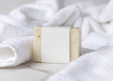 Beyaz banyo havlularının yanında boş kağıt etiketli bej, katı sabun kalıbının bir kopyası. Dikdörtgen şeklinde. Tarafsız arkaplanda, fotokopi alanı ile markasız paketleme, üst görünüm. Kişisel bakım bağlamı