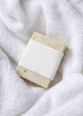 Beyaz banyo terli havlularının üzerinde boş kağıt etiketli bej, katı sabun kalıbının bir kopyası. Tarafsız arkaplanda, fotokopi alanı ile markasız paketleme, üst görünüm. Kişisel bakım bağlamı