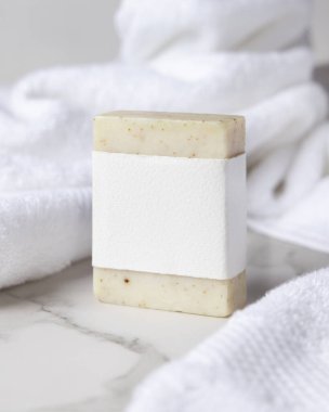 Beyaz banyo havlularının yanında boş kağıt etiketli bej, katı sabun kalıbının bir kopyası. Dikdörtgen şeklinde. Tarafsız arkaplanda, fotokopi alanı ile markasız paketleme, üst görünüm. Kişisel bakım bağlamı