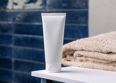 Mavi banyodaki bej banyo havlusunun yanında beyaz, boş plastik kozmetik tüp maketi, kopyalama alanı. Krem, losyon ya da jel ambalajlama. Cilt ürünleri