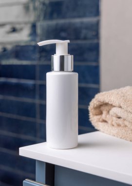 Beyaz pompalı kozmetik şişesi koyu mavi banyoda bej havlunun yanında. Kapatın, kopyalayın. Gümüş metal yakalı boş plastik bir kap. Losyon, krem ya da temizlik malzemesi. Cilt ürünleri