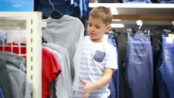 Bonito menino sorridente fica perto de roupas e escolher — Vídeo de Stock