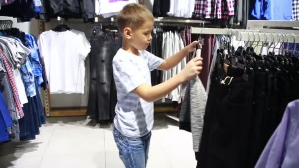 Милый улыбающийся мальчик стоит рядом с одеждой и выбирает — стоковое видео