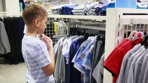 Bonito menino sorridente fica perto de roupas e escolher — Vídeo de Stock