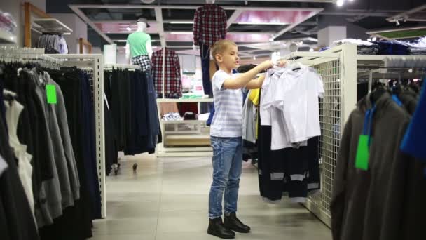 Bonito menino sorridente fica perto de roupas e escolher — Vídeo de Stock